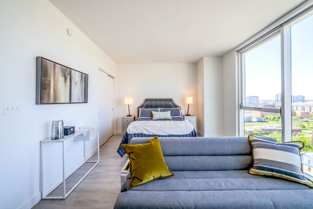 Modern bedroom with a large bed, two nightstands with lamps, a sofa, a wall-mounted TV, and large windows offering a city view.