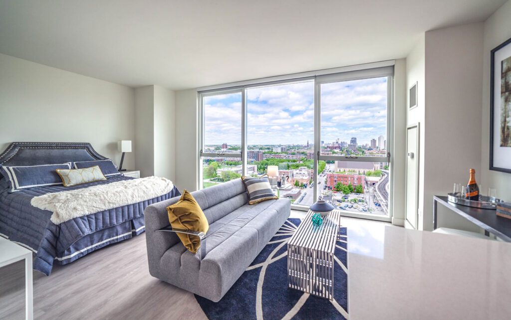 Modern bedroom with large window showing cityscape, featuring a bed, gray sofa with yellow pillow, and a small bar area with a bottle and glasses on a tray.