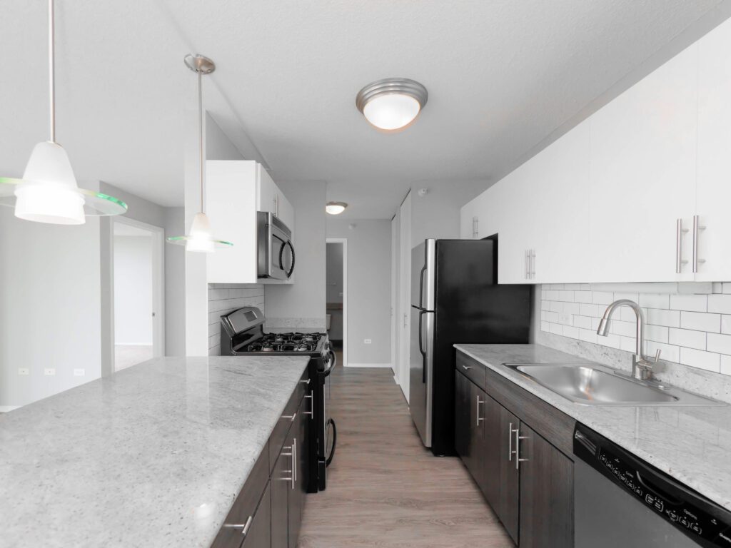 Modern kitchen with white and dark cabinets, stainless steel appliances, and a marble countertop. Features a sink, stove, refrigerator, pendant lights, and a tile backsplash.
