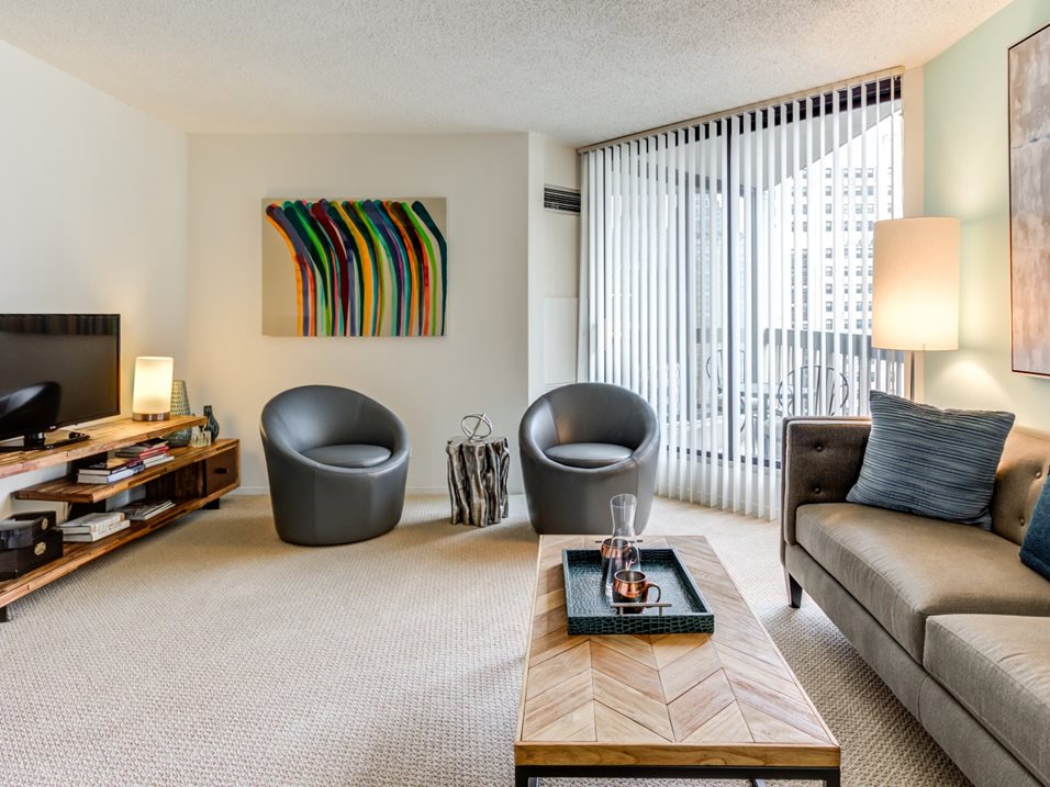 Modern living room with two gray chairs, abstract artwork on the wall, a large sofa, and a wooden coffee table. A TV stands on a low shelf unit, and vertical blinds cover the window.