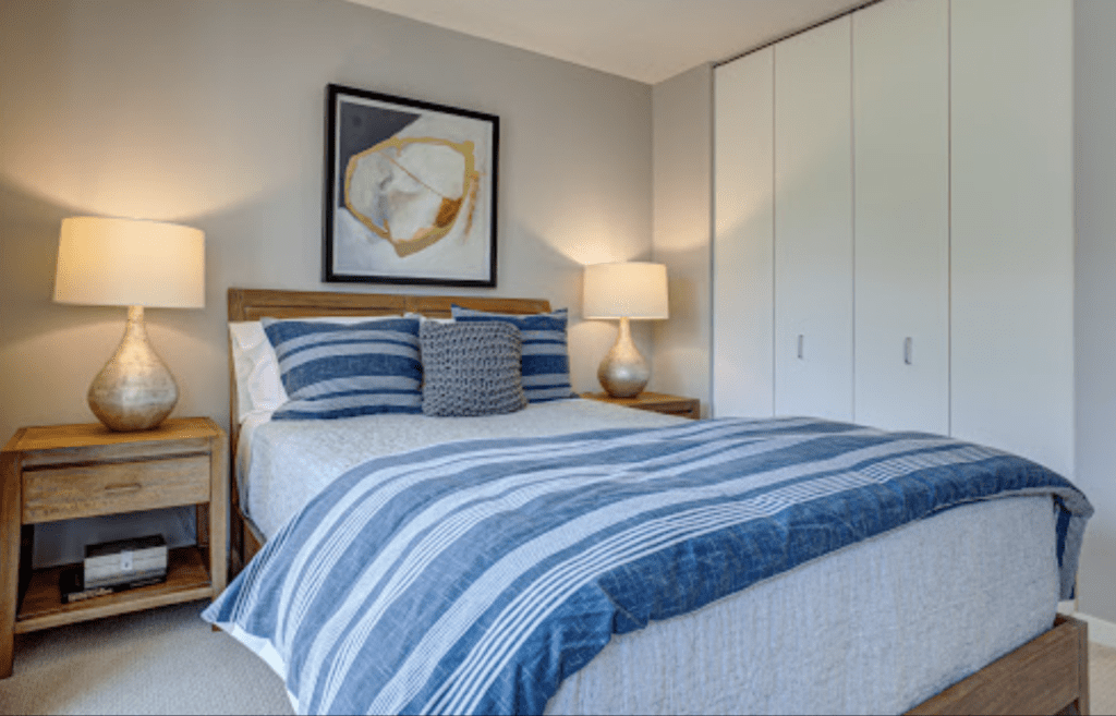 A bedroom with a neatly made bed featuring blue-striped bedding. Two lamps on wooden nightstands flank the bed. A modern abstract painting hangs above the bed. White closet doors are visible.