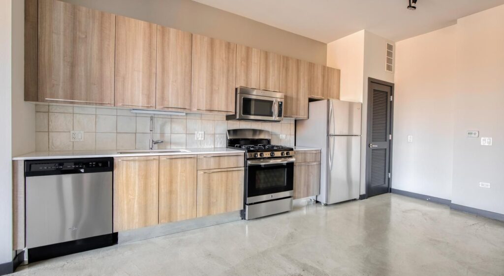 Modern kitchen with light wood cabinets, stainless steel appliances including a refrigerator, stove, microwave, and dishwasher. Neutral-toned walls and flooring.