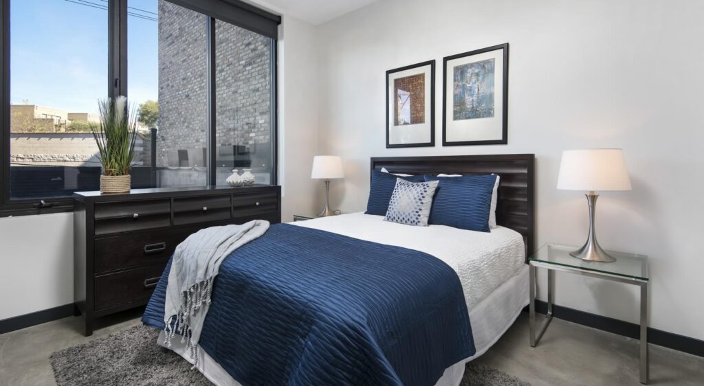 A modern bedroom with a bed featuring blue and white bedding, flanked by two lamps on side tables. A dresser with a plant and art on the walls completes the decor.