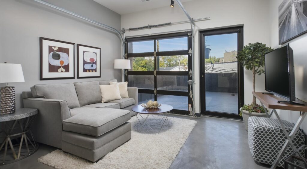 Modern living room with a gray sectional sofa, abstract art on the wall, a TV on a stand, and a glass garage-style door leading outside. Natural light illuminates the space.