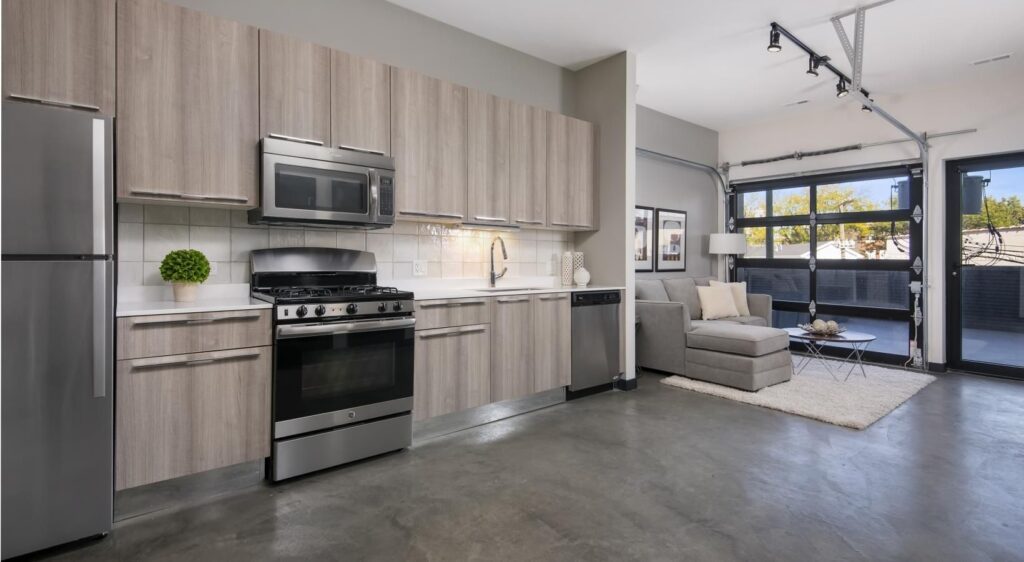 Modern kitchen with wood cabinets, stainless steel appliances, and a concrete floor. An adjacent living area features a sofa, coffee table, and glass doors leading to an outdoor space.