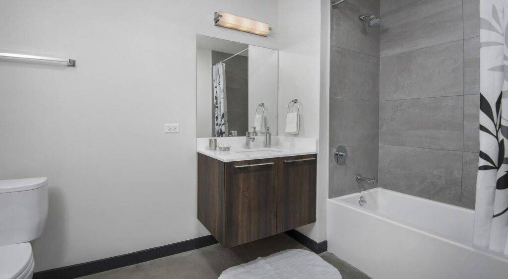 Modern bathroom with a floating vanity, mirror, and wall-mounted light. To the right is a bathtub with a shower and a patterned curtain. The floor is covered with a light gray rug.