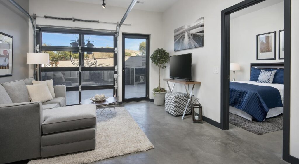 Modern living room with a gray sectional sofa, wall-mounted TV, and open bedroom. Large windows and glass doors provide natural light. Concrete floors and neutral decor create a minimalist style.