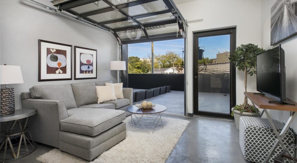 Modern living room with gray sofa, two abstract paintings, a TV, and a glass garage door opening to a patio. Concrete floor and a round rug add to the contemporary style.