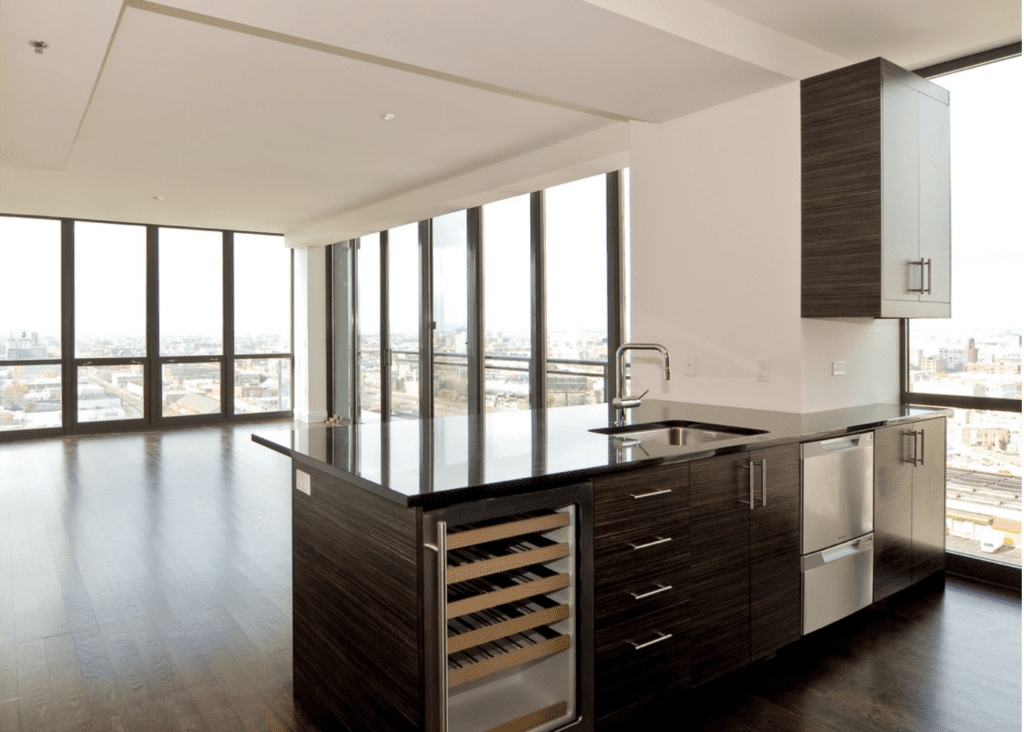 Modern apartment interior with large windows, dark wood floors, and a kitchen featuring dark wood cabinets, a black countertop, and a wine cooler. Cityscape visible through the windows.