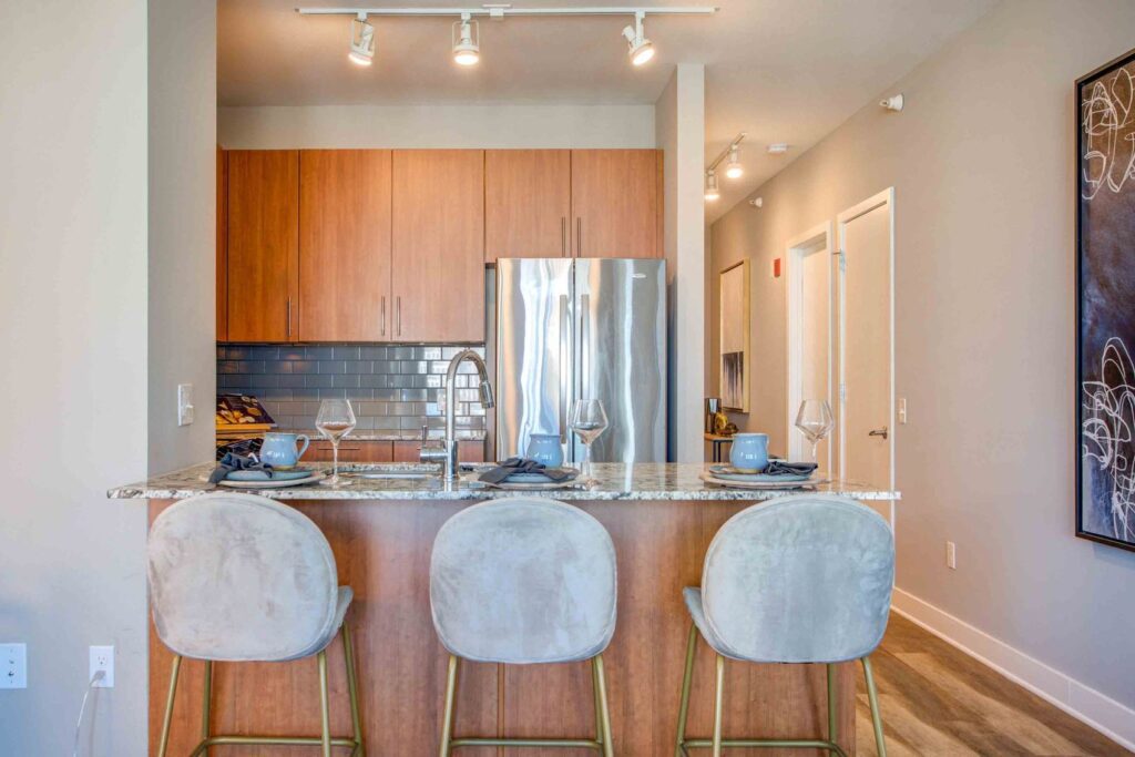 Modern kitchen with wooden cabinets, a marble island, three beige barstools, set with plates and wine glasses, stainless steel appliances, and a large black and white artwork on the right wall.