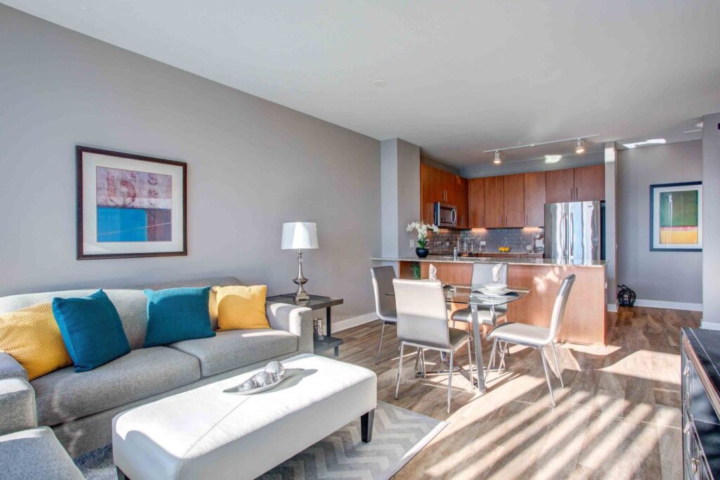 Modern living room and kitchen area with gray sofa, colorful cushions, dining table, wooden flooring, and stainless steel appliances. Wall art and a table lamp add decor.