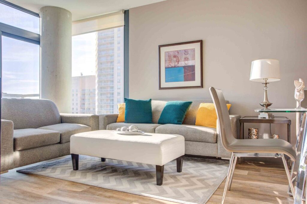 Bright, modern living room with a gray sofa, colorful cushions, a white ottoman, glass table, and large windows showcasing a cityscape.