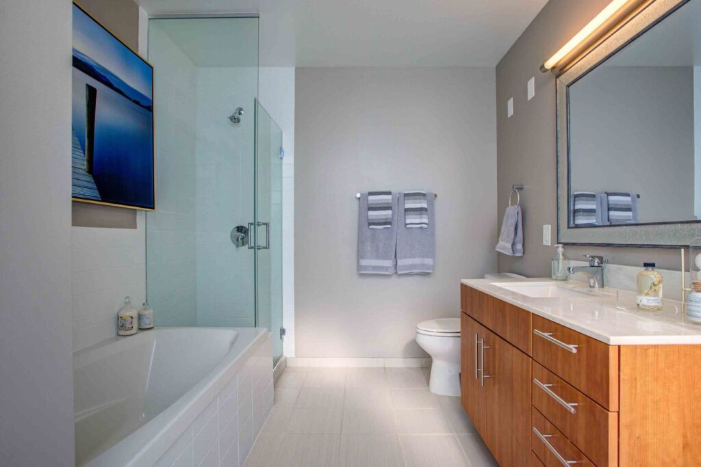 Modern bathroom with a bathtub, glass shower, large mirror, wooden vanity with double sinks, and a towel rack on a gray wall. A framed picture hangs above the tub.