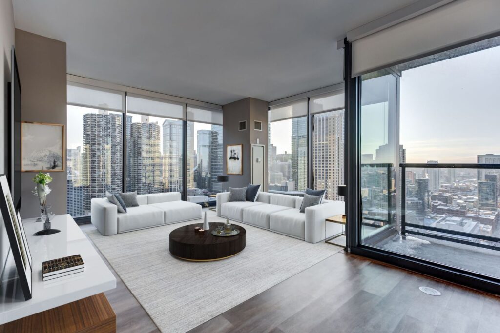 Modern living room with white sofas, a round coffee table, and floor-to-ceiling windows offering a cityscape view with skyscrapers. The room features neutral decor and wooden flooring.