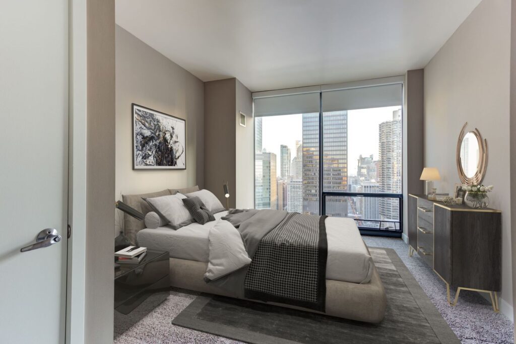 Modern bedroom with a large bed, artwork on the wall, and a dresser. A floor-to-ceiling window reveals a cityscape view with tall buildings in the background.