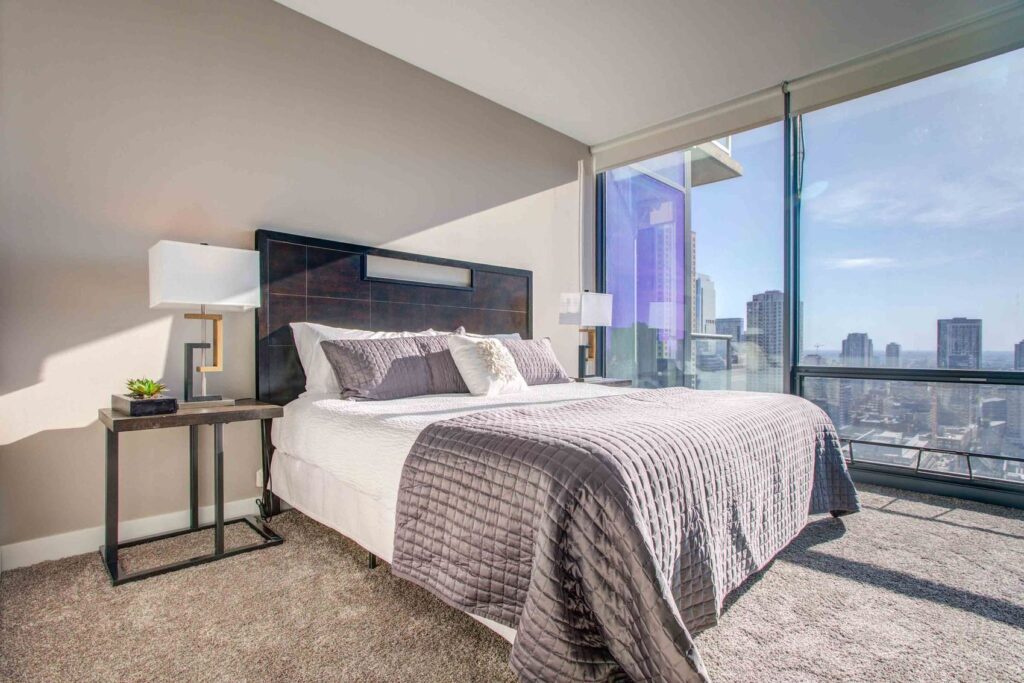 Modern bedroom with a large bed covered in gray bedding, two side tables with lamps, and a floor-to-ceiling window offering a cityscape view.