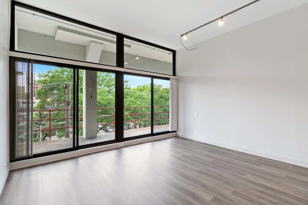 Empty room with large floor-to-ceiling windows, light wood flooring, and a balcony overlooking green trees. Minimalist track lighting along the ceiling.