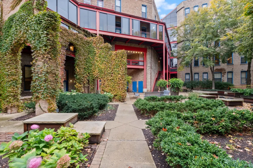 Courtyard with brick buildings, red metal accents, ivy-covered walls, and landscaped garden beds. Benches and potted plants are visible along the pathway.