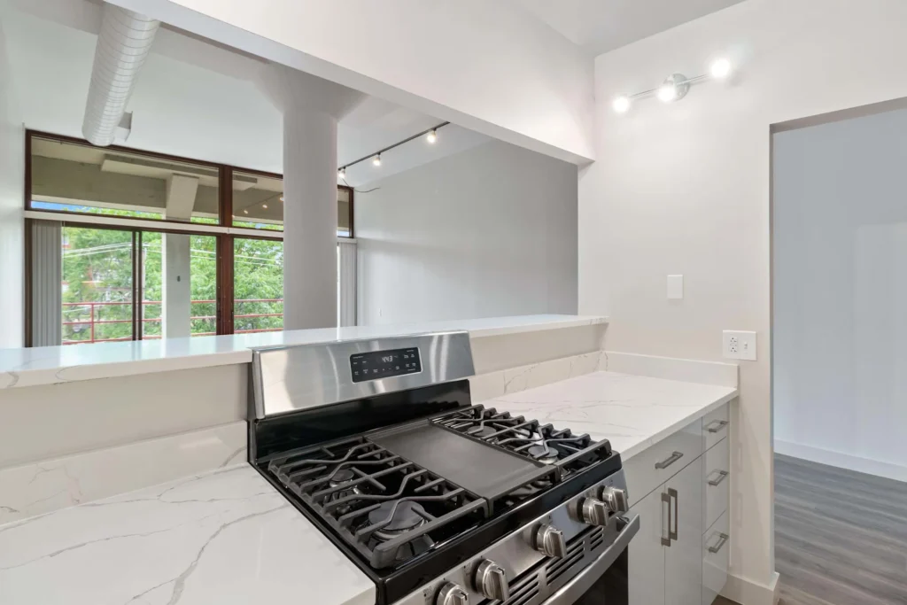 Modern kitchen with a stainless steel gas stove, white marble countertops, and an open layout with large windows in the background.