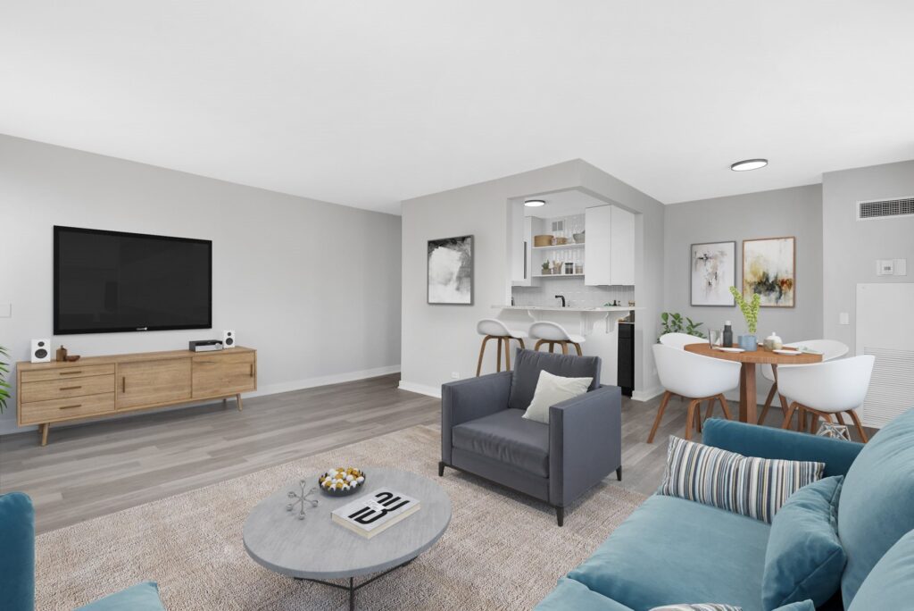 Modern living room with gray couch, armchair, and rug. A flat-screen TV is on a wooden console. The dining area features a round table and chairs, with a small open kitchen in the background.