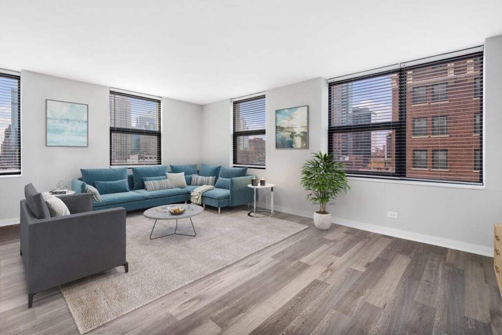 Modern living room with a teal sectional sofa, gray armchair, and round coffee table on a rug. Large windows offer a city view. Artwork and a potted plant decorate the space.