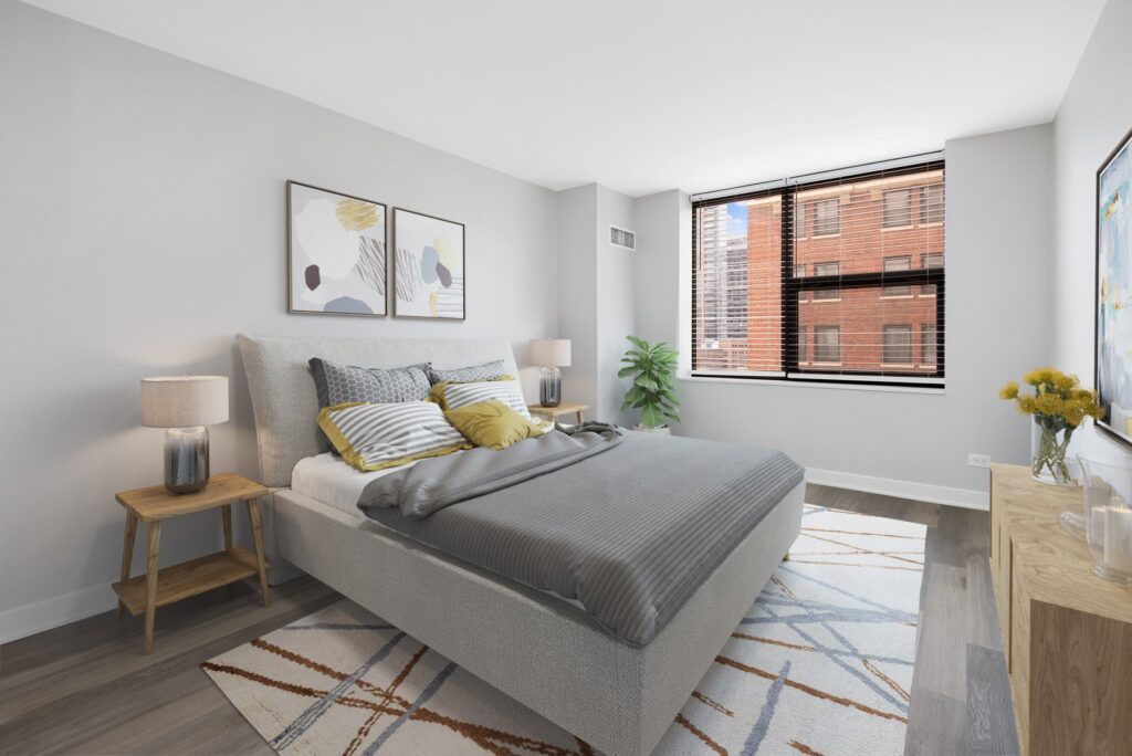 A modern bedroom with a gray bed, geometric artwork, two bedside tables with lamps, a wooden dresser, and a window with a city view. A rug covers part of the wooden floor.