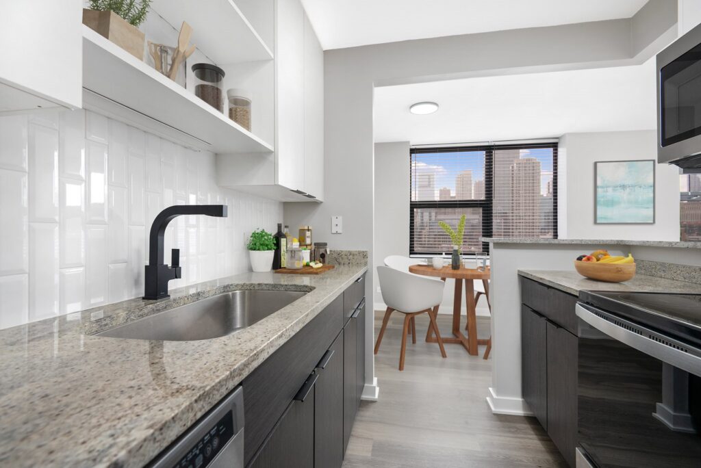 Modern kitchen with granite countertops, a black faucet, and stainless steel appliances. Dining area with wooden table and white chairs near windows showing a cityscape.