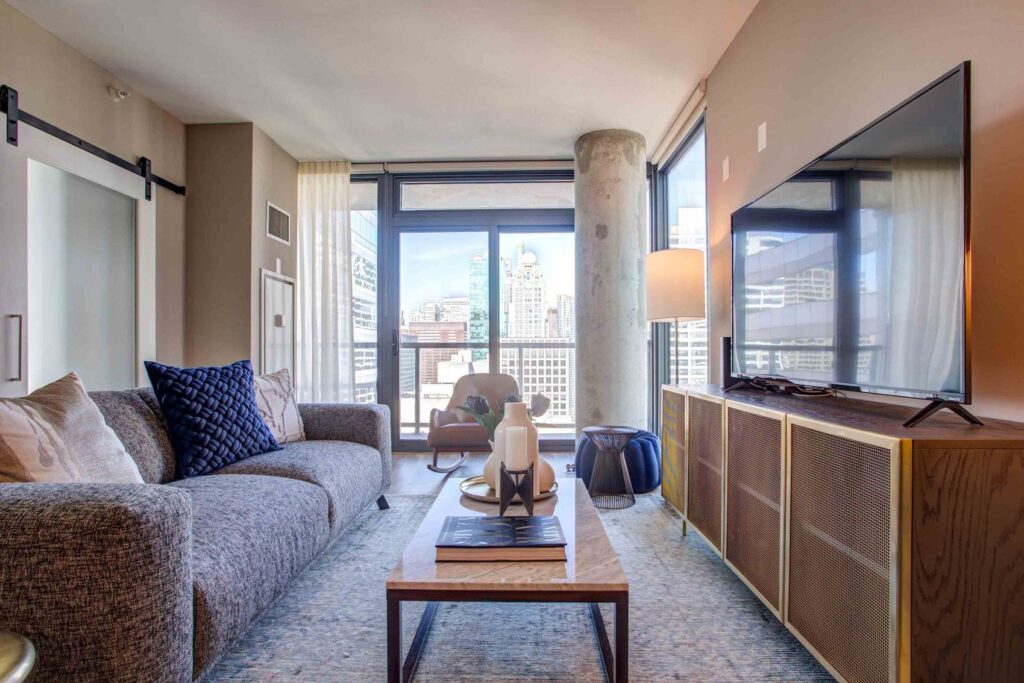Modern living room with a gray couch, blue pillows, a wooden coffee table, large TV, and floor-to-ceiling windows showing a cityscape.