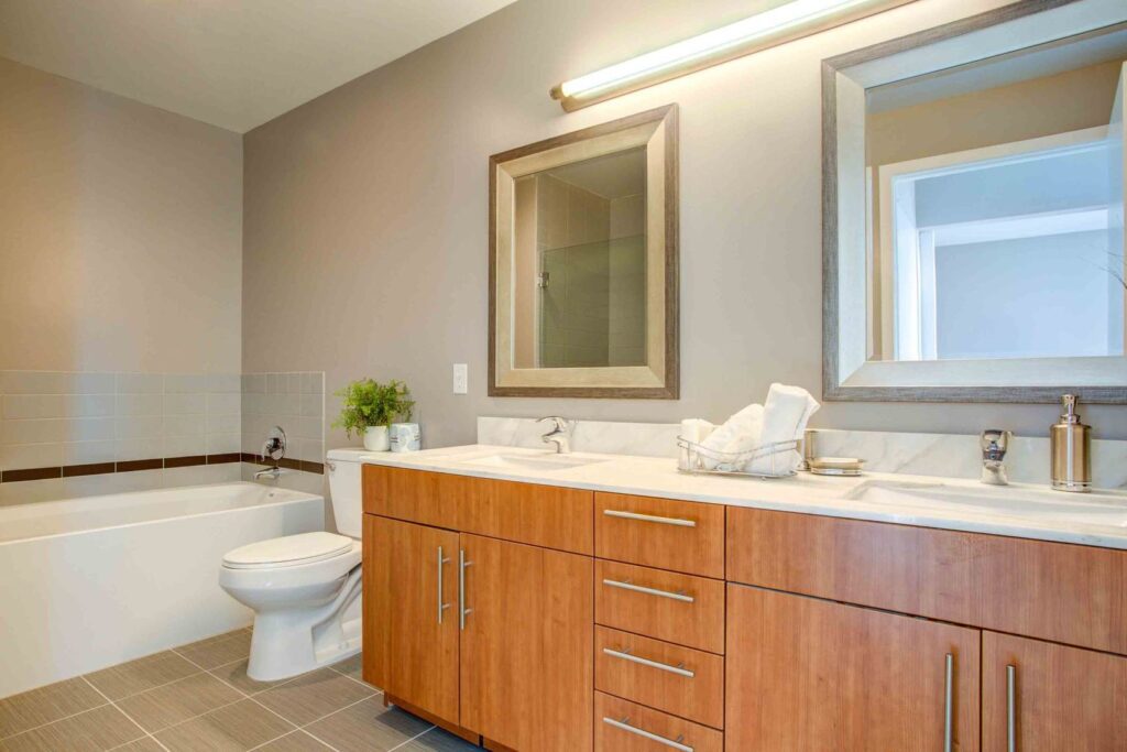 Modern bathroom with wooden vanity featuring dual sinks, a large mirror, a towel holder, and a plant. A bathtub is positioned near the vanity, with neutral-toned walls and tiles.