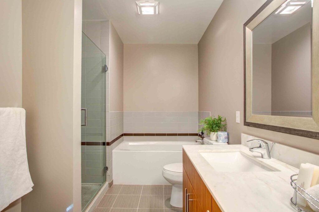 Modern bathroom with a glass-enclosed shower, bathtub, white sink on a wooden cabinet, large mirror, and neutral walls. Towels hang on a rack; a plant is placed near the sink.