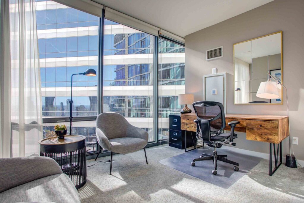 Modern office space with a desk, chair, and lamp near a large window. A round side table and chair are in the foreground, and a building is visible outside the window.