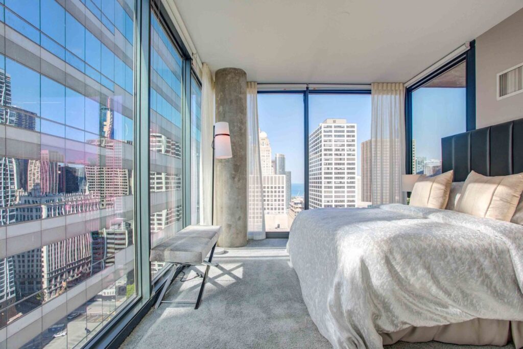 Modern bedroom with large windows offering a cityscape view. Room features a bed with white bedding, a bench, and curtains framing the windows. Sunlit high-rise buildings are visible outside.