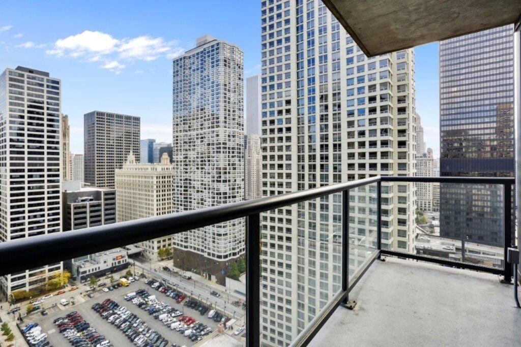 View from a high-rise balcony overlooking a cityscape with numerous tall buildings and a parking lot below. Clear blue sky in the background.
