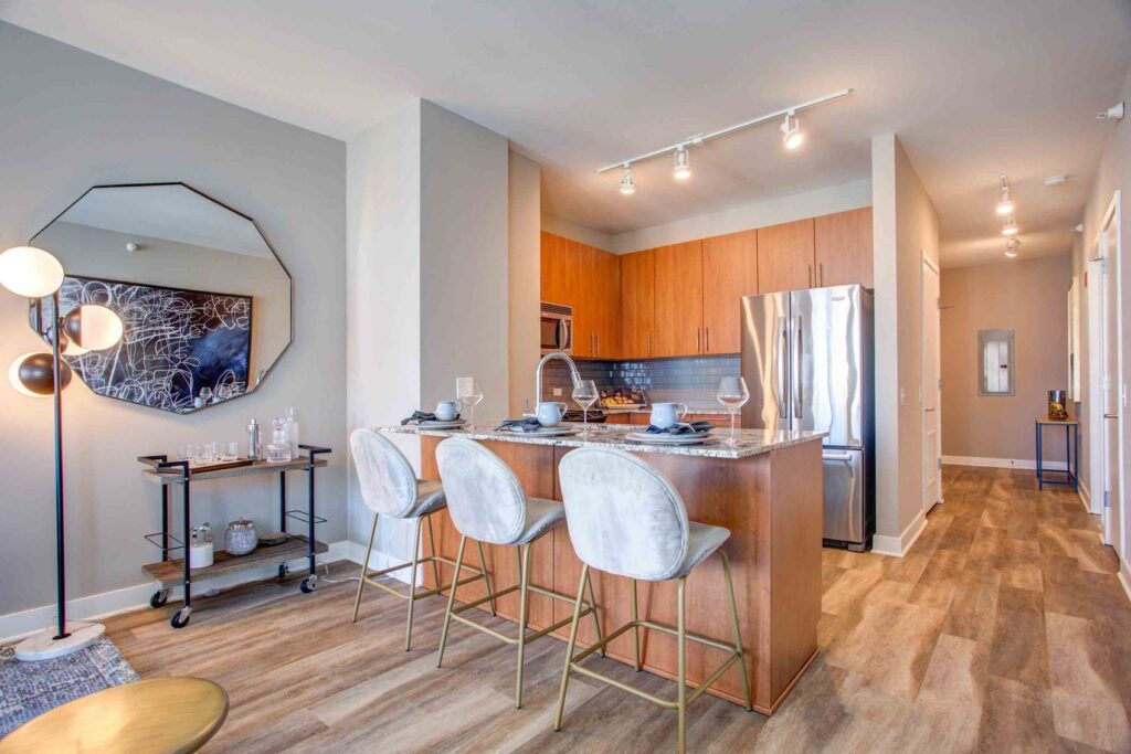 Modern kitchen with wooden cabinets, stainless steel appliances, and three gray chairs at an island. A bar cart and a floor lamp with three bulbs are on the left. A hallway is visible on the right.