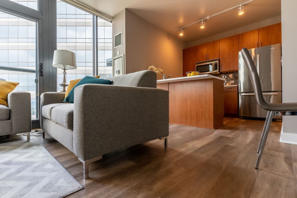 Modern living room and kitchen with gray sofa, wooden cabinets, stainless steel appliances, and large windows.
