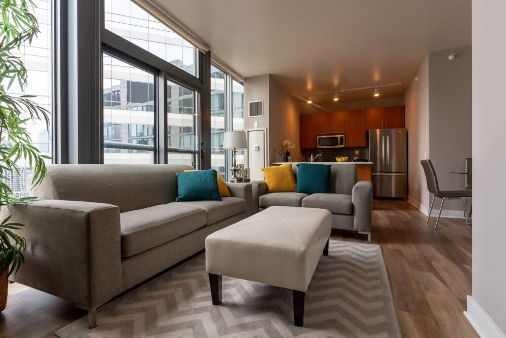 Modern living room with a gray sofa, teal and yellow cushions, a white ottoman, zigzag rug, and a view of a kitchen with stainless steel appliances. Large windows and wooden flooring are visible.
