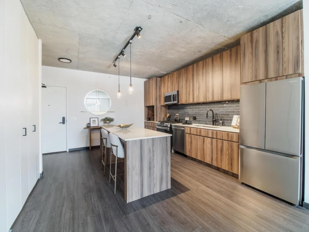 Modern kitchen with wood cabinetry, stainless steel appliances, island with stools, and gray flooring. A round mirror and pendant lights decorate the space.