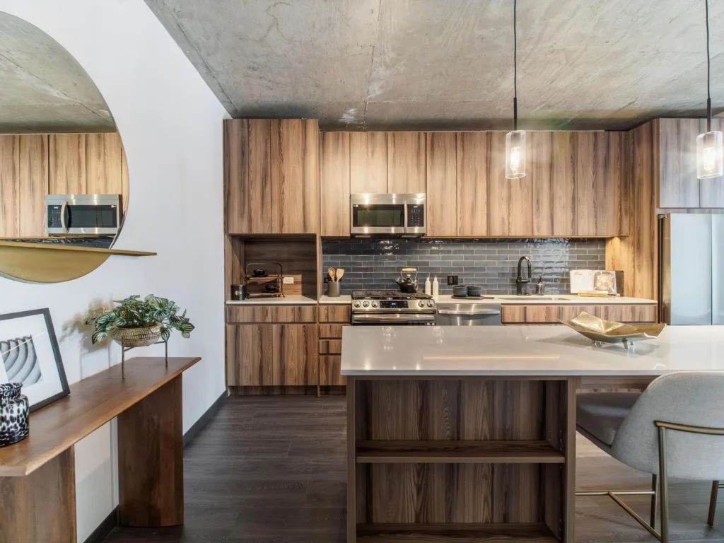 Modern kitchen with wooden cabinets, dark backsplash, and white island counter. Includes stainless steel appliances, pendant lights, and decorative mirror and plant on a side table.