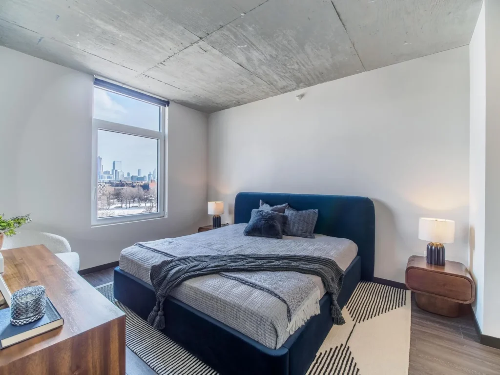 Modern bedroom with a large bed, gray bedding, and blue accents. Two bedside tables with lamps, a view of a city skyline through the window, and a wooden dresser complete the decor.