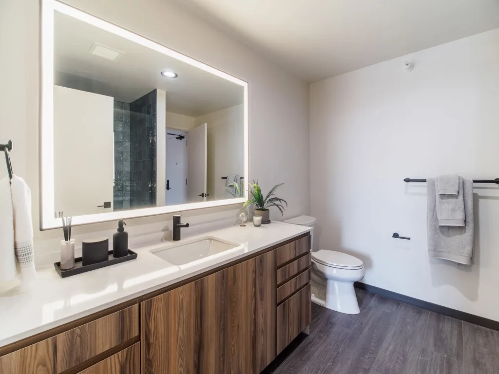 Modern bathroom with a large illuminated mirror, wooden vanity, and white countertop. Includes a sink, faucet, and various toiletries. A towel hangs beside a toilet on the right.