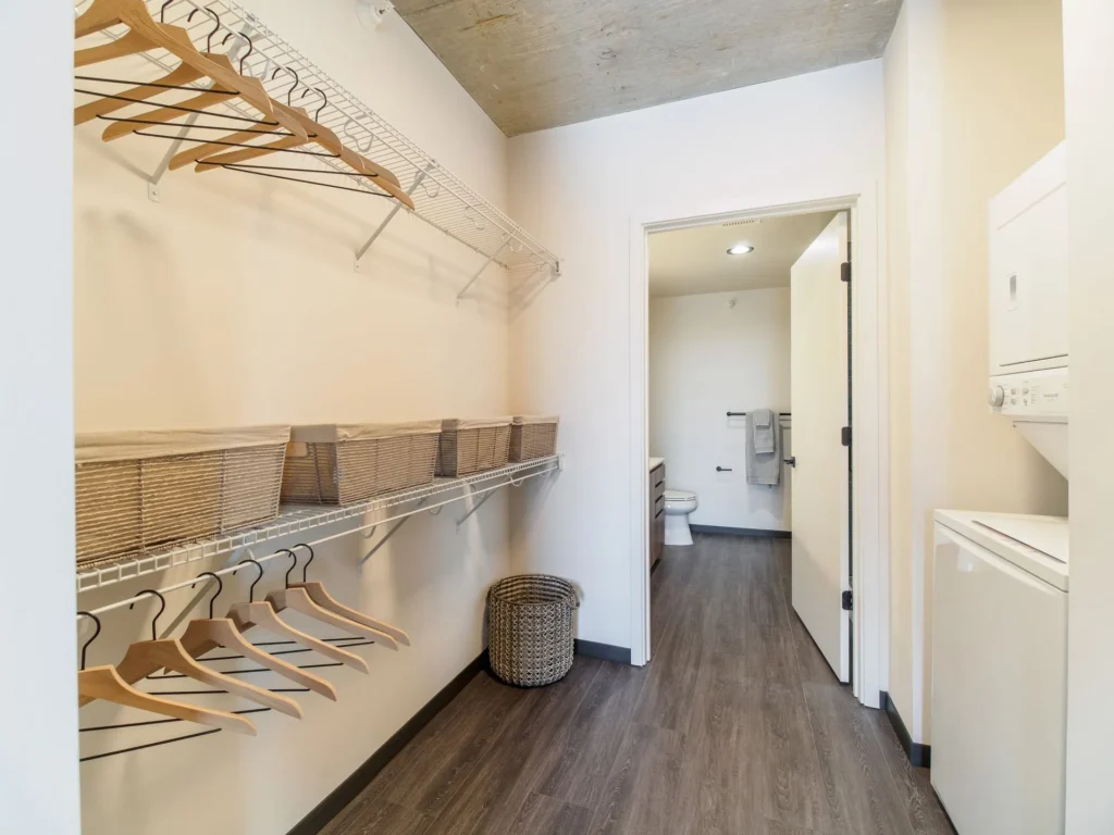 A laundry room with a stackable washer and dryer, wire shelves with baskets, hangers, and a wicker basket. The room has a wooden floor and leads to a bathroom.