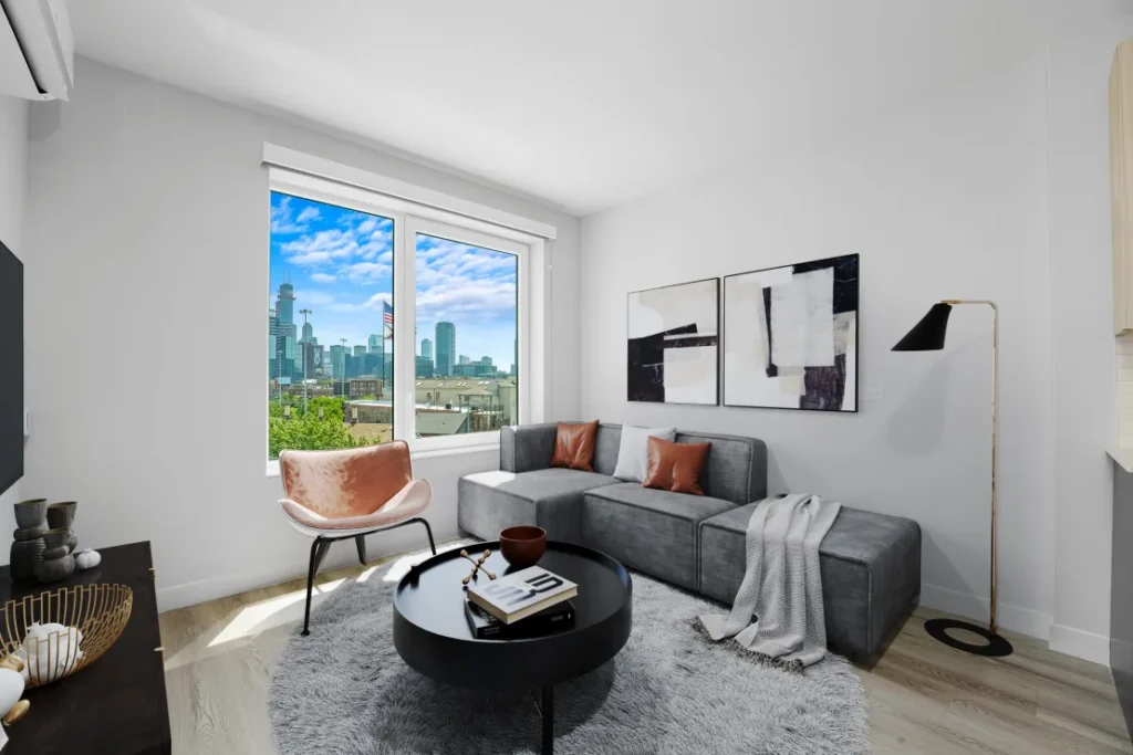 Modern living room with a large window showing a cityscape. Features a gray sectional sofa, black round coffee table, pink chair, and abstract wall art. Light wood flooring and neutral decor.