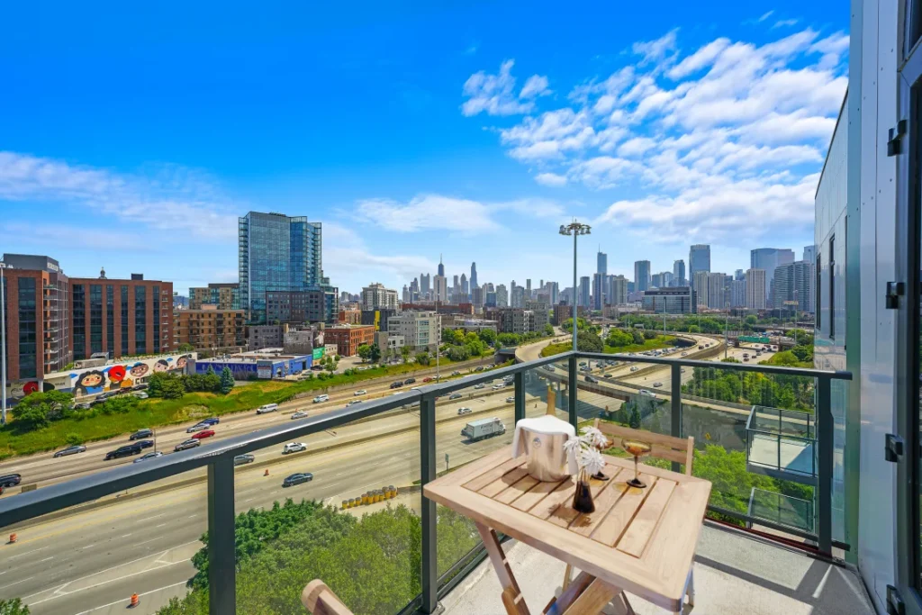 View from a balcony overlooking a highway with multiple lanes, surrounded by urban buildings and a city skyline in the background under a blue sky with scattered clouds.