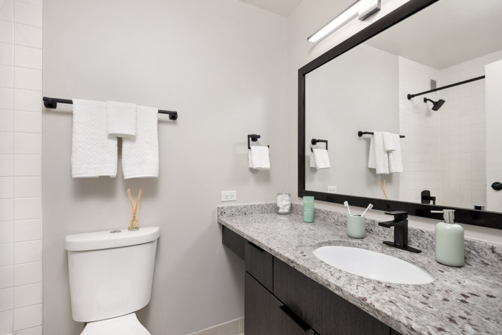 Modern bathroom with a white toilet, granite countertop with a sink, black fixtures, wall-mounted mirror, and a towel rack holding white towels.