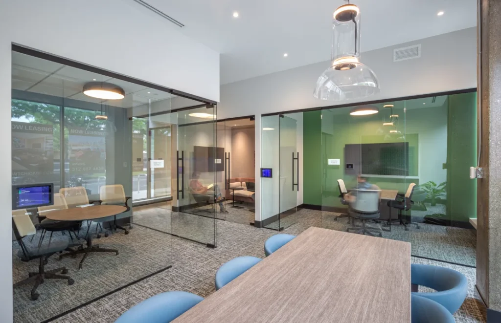 Modern office space with glass-walled meeting rooms, pendant lights, a long table with blue chairs, and two smaller round tables with chairs.