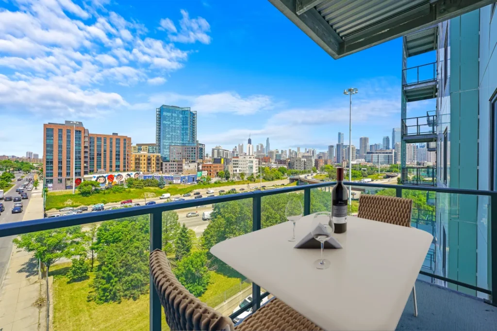 View from a balcony with a table set for two, overlooking a cityscape with a clear blue sky. A bottle of wine and a glass sit on the table.