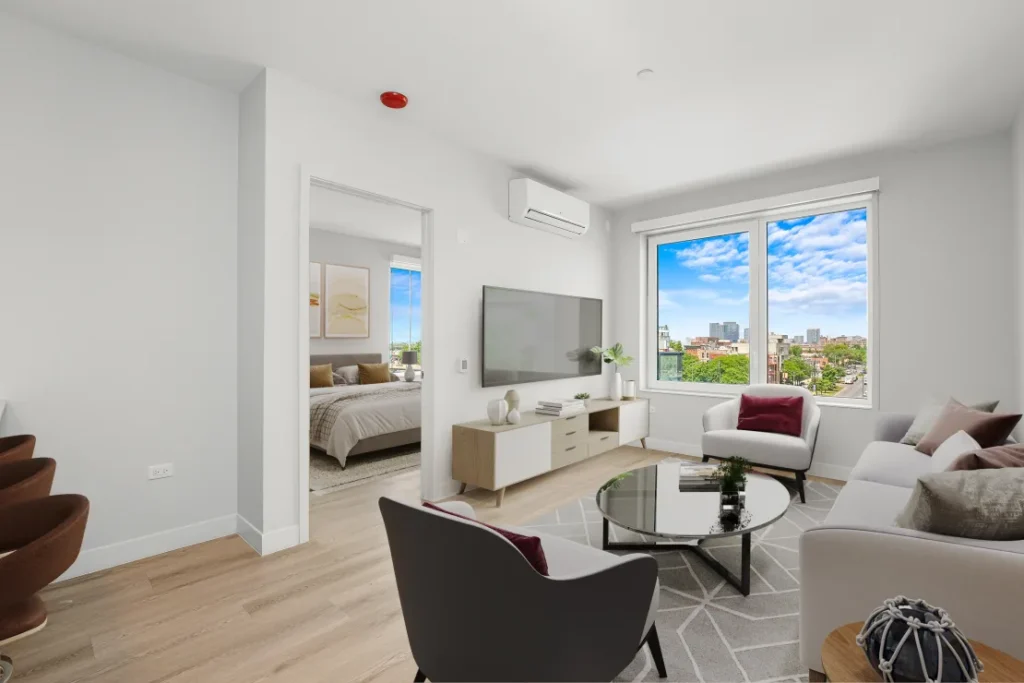 Modern living room with white walls, featuring a sofa, armchairs, and a glass coffee table. A large window offers a city view. A wall-mounted TV and air conditioning unit are present. Bedroom visible.