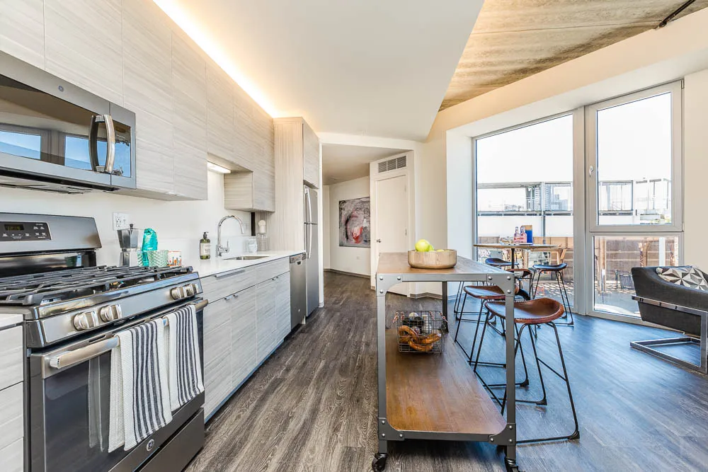 Modern kitchen with stainless steel appliances, light wood cabinetry, and a kitchen island. Floor-to-ceiling windows offer a city view. A small dining area features tall chairs with a round table.