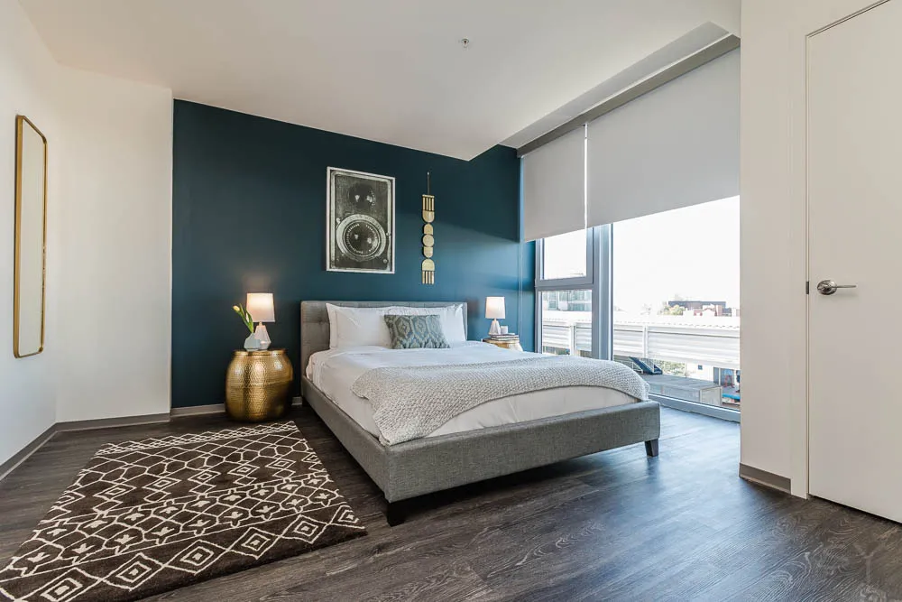 Modern bedroom with a blue accent wall, a large bed, patterned rug, and a small round table with a lamp. Large window with blinds on the right, and a closed door on the left.