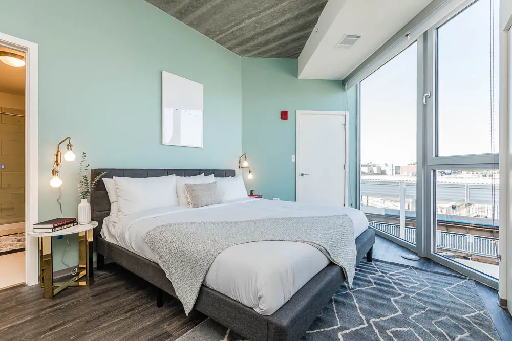 A well-lit bedroom with a large bed, green walls, and a small side table with lamps. Large windows offer a city view. A geometric-patterned rug covers part of the wooden floor.